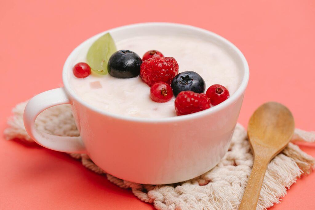 "Tigela branca com creme de cupuaçu suave, coberto por frutas frescas, incluindo mirtilos, framboesas, groselhas vermelhas e uma fatia de uva verde. Ao lado, uma colher de madeira sobre um suporte de crochê em tom bege, tudo em um fundo cor-de-rosa suave."